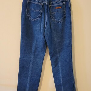 May include: Dark blue denim jeans with a classic five-pocket design. The jeans have a high waist and a straight leg. The back pockets feature a decorative stitch. The brand name is visible on a small patch.