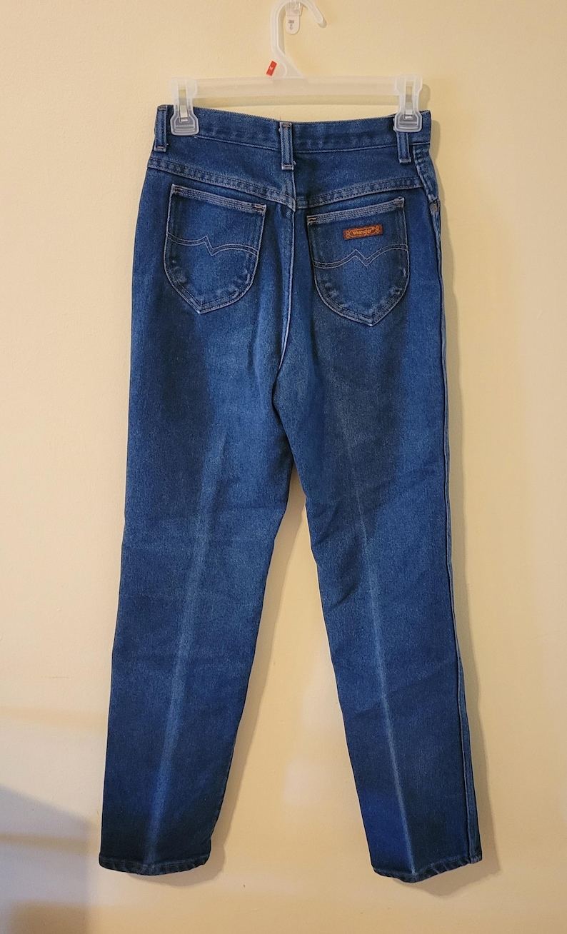 May include: Dark blue denim jeans with a classic five-pocket design. The jeans have a high waist and a straight leg. The back pockets feature a decorative stitch. The brand name is visible on a small patch.