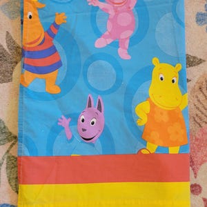 May include: A blue bed sheet with cartoon characters in orange, pink, purple, and yellow. The sheet has a blue background with large blue circles. The bottom of the sheet has horizontal stripes of red and yellow.