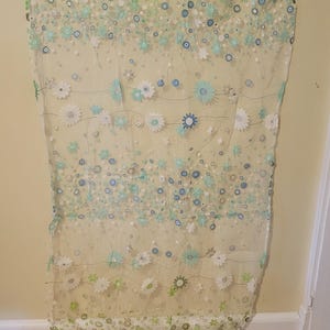 Può includere: Pannello tenda trasparente con motivo floreale. Il pannello è decorato con applicazioni e paillettes di fiori bianchi, verdi e blu. La tenda è appesa a un'appendiabiti nero contro un muro giallo chiaro.