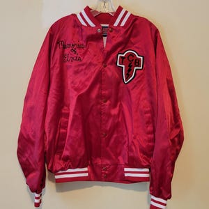 Puede incluir: Chaqueta de satén roja con rayas blancas en el cuello, los puños y el dobladillo. La chaqueta presenta el texto bordado "Memories of Elvis" y un parche con "TCB" y un diseño de rayo. La chaqueta está abotonada y colgada de una percha.