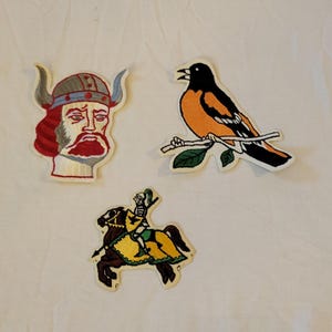 May include: Three embroidered patches: a Viking head with a horned helmet, an orange and black bird perched on a branch, and a knight on horseback. The patches have a cream-colored border and are on a white background.