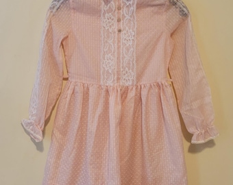 May include: A light pink long-sleeve dress featuring a subtle vertical striped pattern with white swiss dot texture. The dress includes a pointed collar, a front button placket, and white floral lace trim along the shoulders and hem.