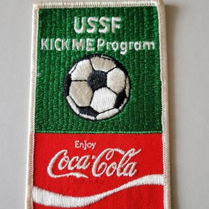 Puede incluir: Parche bordado con un balón de fútbol y el texto "USSF KICK ME Program" sobre un logotipo rojo de Coca-Cola. El parche tiene una sección superior verde y una inferior roja, con costuras blancas.