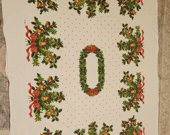 May include: A white rectangular tablecloth featuring a repeating pattern of green holly leaves, red ribbons, and gold bells. The center displays an oval wreath of holly and red ribbon, surrounded by small red dots across the fabric surface.