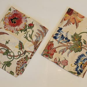 May include: Two folded floral fabric pieces with a cream background. The fabric features large red, pink, and blue flowers with green leaves and brown stems. The design is repeated across both pieces.