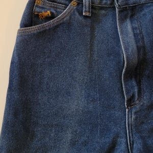 May include: Close-up of blue denim jeans. The jeans have a small pocket with a horse logo, a button closure, and a classic five-pocket design. The denim has a medium wash and visible stitching.