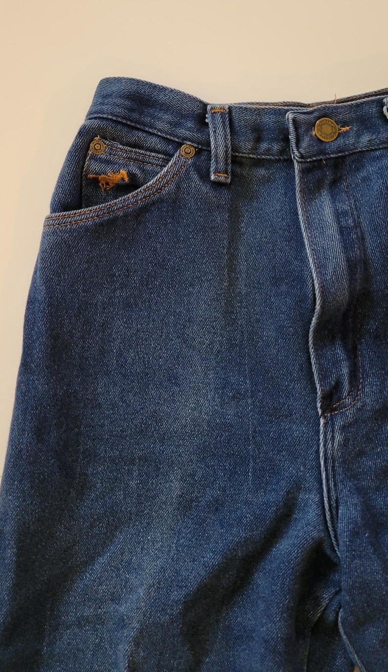 May include: Close-up of blue denim jeans. The jeans have a small pocket with a horse logo, a button closure, and a classic five-pocket design. The denim has a medium wash and visible stitching.