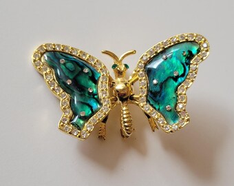 May include: A gold-toned butterfly brooch with iridescent teal and green wings. The wings are outlined with clear crystals. The body and antennae are gold, and the eyes are small green stones. A decorative jewelry item.
