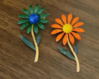 May include: Two flower-shaped brooches with long stems and leaves. One brooch features green petals with a blue center, while the other has orange petals with a yellow center. Both items have gold-toned stems and green leaves attached to the base.