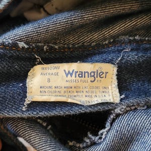 May include: Close-up of blue denim jeans with a Wrangler label. The label indicates size 8 and washing instructions. The jeans are made of denim fabric, showing signs of wear and tear.
