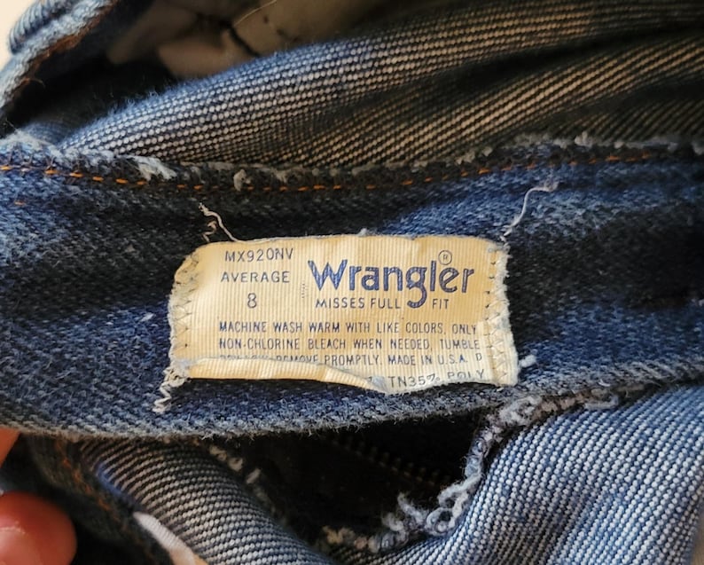 May include: Close-up of blue denim jeans with a Wrangler label. The label indicates size 8 and washing instructions. The jeans are made of denim fabric, showing signs of wear and tear.