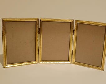 May include: A triptych photo frame featuring three rectangular gold-toned metal frames connected by hinges. Each frame has a textured, beaded border design and contains a plain, light brown backing board, allowing for the display of three photos.