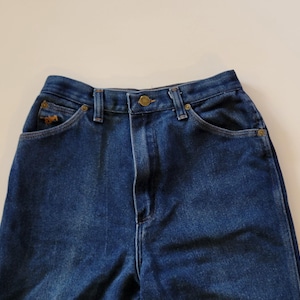May include: Close-up of blue denim jeans. The jeans feature a high waist, button closure, and pockets. A small embroidered horse logo is visible on the front pocket. The jeans are made of a sturdy denim material.