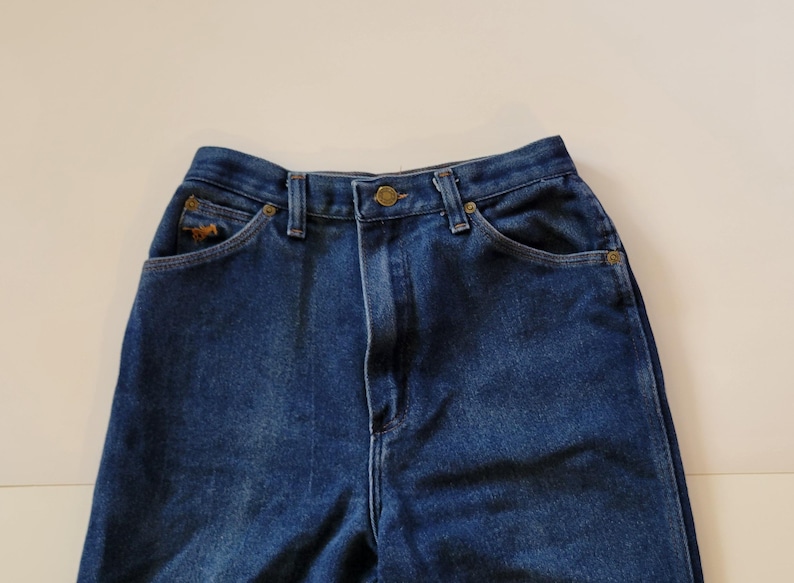 May include: Close-up of blue denim jeans. The jeans feature a high waist, button closure, and pockets. A small embroidered horse logo is visible on the front pocket. The jeans are made of a sturdy denim material.