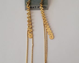 May include: A pair of gold-tone drop earrings attached to a gray display card labeled FASHION EARRINGS and 18 K.T. PLATED. Each earring features a vertical wavy metal strip, a thin chain, and a small smooth gold-tone bead at the bottom end.
