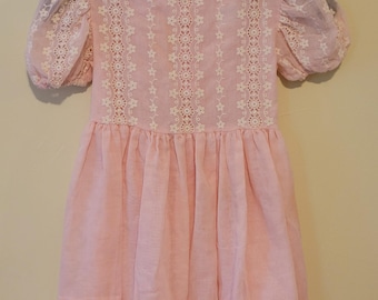 May include: A light pink dress featuring short puff sleeves and a round neckline. The bodice is decorated with vertical panels of white floral lace trim. The skirt has a gathered waistline and a ruffled hem, creating a layered appearance.