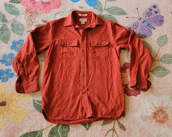 May include: A rust-colored long-sleeve shirt with two chest pockets and a button-down front. The shirt has a collared neckline and buttoned cuffs. The shirt is made of a soft, textured fabric.