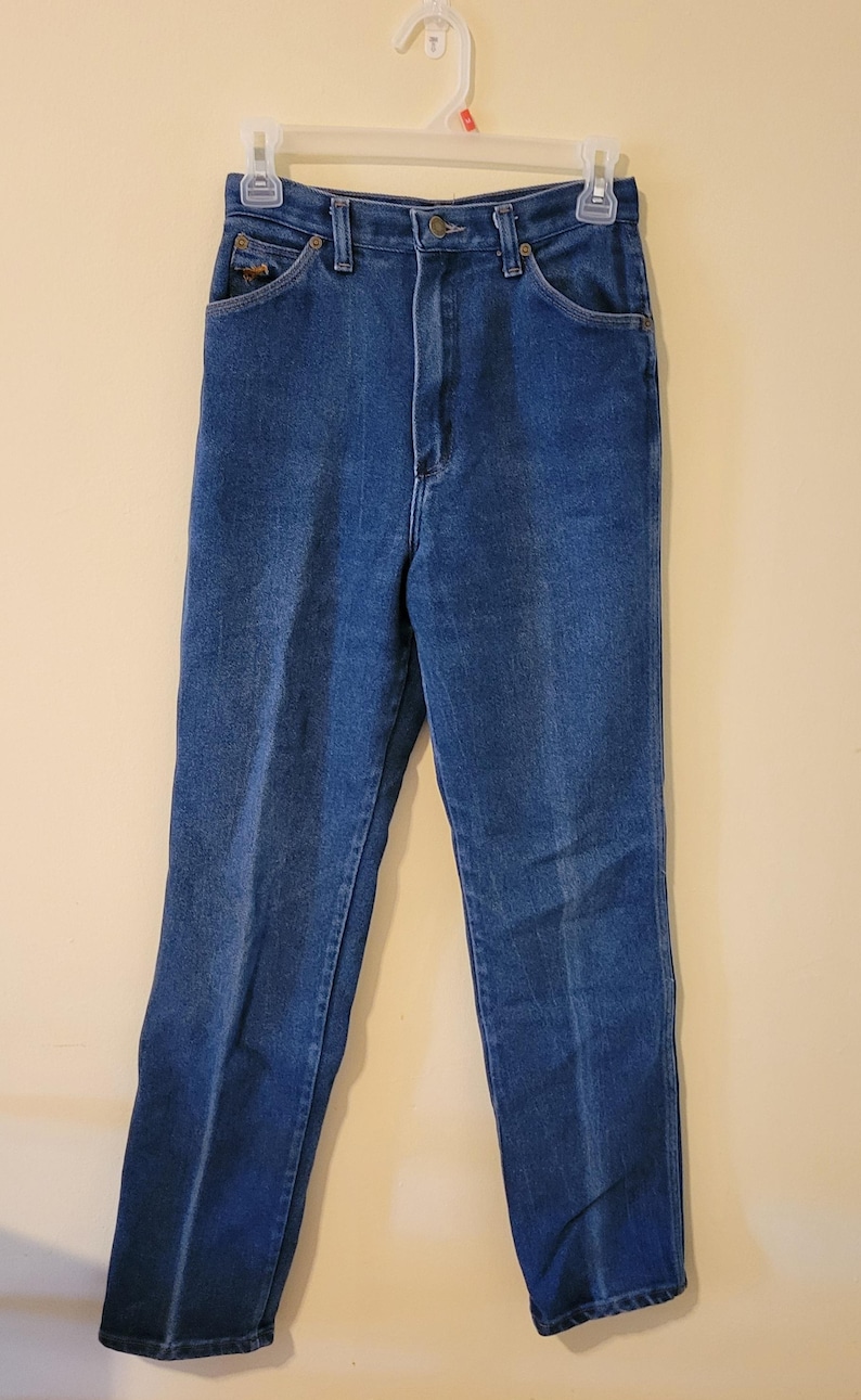 May include: A pair of blue denim jeans hanging on a white hanger. The jeans feature a classic five-pocket design, a button and zip fastening, and a straight leg cut. The denim is a medium wash with subtle fading.