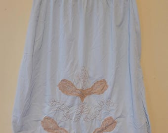 May include: A light blue half slip featuring a beige lace floral applique centered above a scalloped beige lace hem. The garment has an elastic waistband and is displayed on a clear plastic hanger with a yellow size tag attached to the hook.