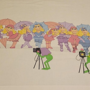 Op de afbeelding: Een witte stof met een herhalend patroon van Miss Piggy-figuren in diverse roze, paarse en blauwe outfits met bijpassende paraplu's, afgewisseld met groene Kermit de Kikker-figuren die camera's op zwarte statieven bedienen.
