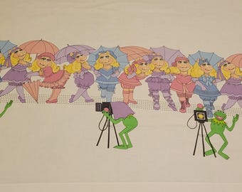 May include: A white fabric featuring a repeating pattern of Miss Piggy characters in various pink, purple, and blue outfits with matching umbrellas, interspersed with green Kermit the Frog figures operating vintage cameras on black tripods.