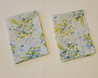 May include: Two rectangular fabric napkins featuring a light blue background with a repeating pattern of small blue and white flowers and green stems. The floral design is arranged in clusters across the surface of both folded textile pieces.