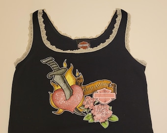 May include: A black tank top with light gray lace trim along the neckline and armholes. The front features a graphic of a dagger piercing a pink heart surrounded by yellow flames, pink flowers, and a banner with the text Harley-Davidson.