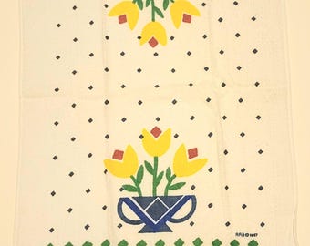 May include: A white kitchen towel featuring two blue vases with yellow flowers and green stems. The design includes a pattern of small black dots and horizontal bands of green, yellow, and red diamonds. The text KITCHEN TOWEL and RABO 1987 are printed.