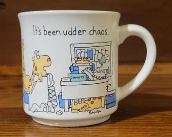 May include: A white ceramic mug featuring a cartoon illustration of cows in an office setting with the text It's been udder chaos. The design includes a cow at a desk with a donut box, a telephone, and a bulletin board, with the artist signature Boynton.