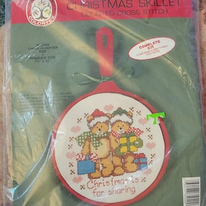 May include: A Colortex Christmas Skillet counted cross stitch kit in a clear plastic package. The kit includes a red molded skillet frame, white Aida cloth, embroidery floss, needle, and ribbon. The design features two teddy bears with gifts.