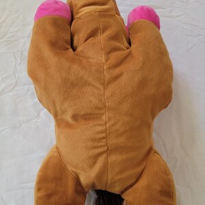 Puede incluir: Un caballo de peluche en posici&oacute;n tumbada. El caballo es principalmente marr&oacute;n con cascos rosas y una nariz rosa. Tiene una melena y cola marrones. El juguete est&aacute; hecho de material suave y afelpado.