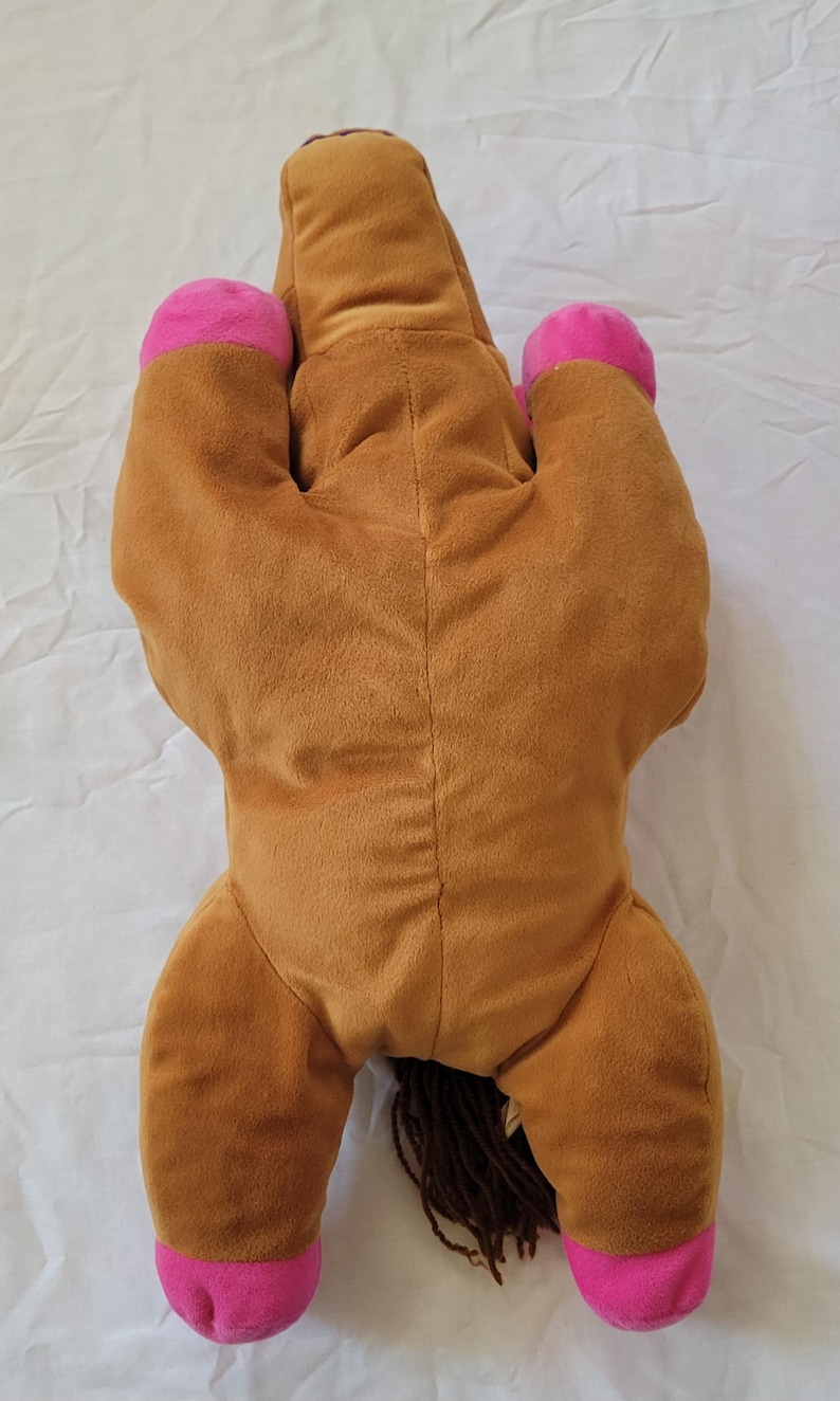 Puede incluir: Un caballo de peluche en posici&oacute;n tumbada. El caballo es principalmente marr&oacute;n con cascos rosas y una nariz rosa. Tiene una melena y cola marrones. El juguete est&aacute; hecho de material suave y afelpado.