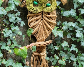 May include: A brown macrame owl wall hanging with green yarn eyes. The owl is hanging on a wall covered in green ivy.