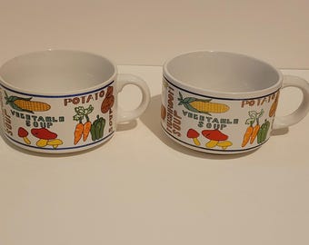 May include: Two white ceramic soup mugs featuring colorful illustrations of vegetables including corn, carrots, and mushrooms. The text "VEGETABLE SOUP" and "POTATO SOUP" is printed on the sides in brown and green lettering with blue trim.