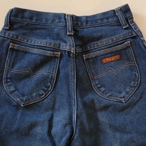 May include: A close-up of the rear of blue denim jeans. The jeans feature two back pockets with a distinctive wave-like stitch. A small brown patch with the "Wrangler" brand name is on the right pocket. The jeans are made from a robust denim fabric.