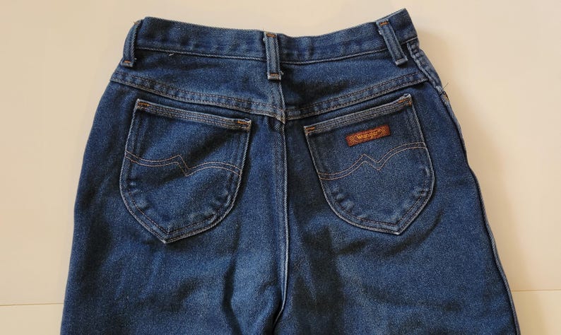 May include: A close-up of the rear of blue denim jeans. The jeans feature two back pockets with a distinctive wave-like stitch. A small brown patch with the "Wrangler" brand name is on the right pocket. The jeans are made from a robust denim fabric.
