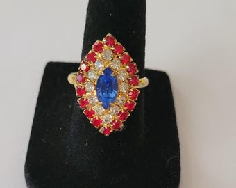 May include: A gold-toned ring featuring a marquise-shaped blue center stone, surrounded by a halo of clear white stones and an outer border of small red stones. The ring has a pointed, elongated design that covers the top of the finger.