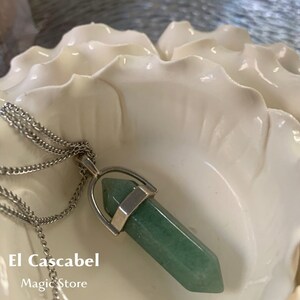 May include: A green gemstone pendant necklace with a silver chain. The pendant is a hexagonal shape. The necklace is displayed in a white, shell-shaped dish. The text "El Cascabel Magic Store" is visible.