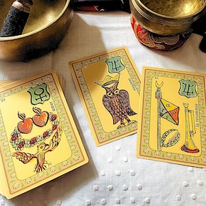 Puede incluir: Cartas de tarot con ilustraciones, incluyendo un búho, un pájaro y otros símbolos. Las cartas están sobre una superficie blanca, con un cuenco tibetano de latón y una maza de madera en el fondo.