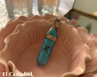 Op de afbeelding: Een turquoise hangerketting met een zilveren ketting. De hanger heeft een zeshoekige vorm met een goudkleurige metalen band. De ketting wordt getoond in een roze, bloemvormige schaal. De tekst "El Cascabel Magic Store" staat onderaan.