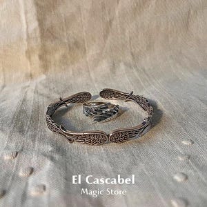 May include: A silver ring and bracelet set featuring a wing design. The ring is open and the bracelet is a bangle style. The set is on a beige fabric background. El Cascabel Magic Store is printed below the set.