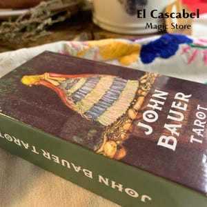 Puede incluir: Una caja verde con el texto "John Bauer Tarot" en la parte delantera y trasera. La caja presenta una ilustración colorida de una mujer con un vestido largo y fluido de pie en un bosque.