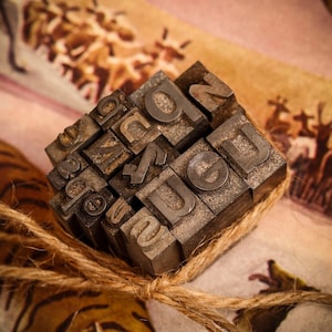 May include: A collection of vintage metal letterpress printing blocks, arranged in a random pattern, tied together with twine. The blocks are made of metal and have a worn, aged look. The letters on the blocks are a mix of uppercase and lowercase letters.