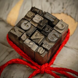 Könnte beinhalten: Ein rotes Band um eine Sammlung von alten Metall-Letterpress-Druckblöcken gebunden. Die Blöcke sind in Würfelform angeordnet und zeigen verschiedene Buchstaben und Zahlen.