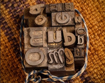 May include: A collection of vintage metal letterpress printing blocks, tied together with twine, on a brown and purple woven fabric background.