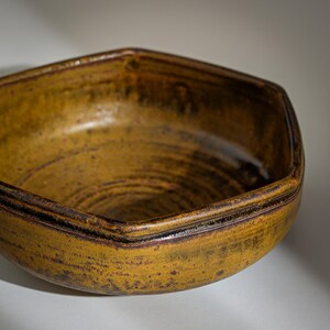 May include: A hexagonal ceramic bowl with a dark brown glaze and a speckled yellow finish. The bowl has a wide, shallow shape and a slightly rounded bottom.