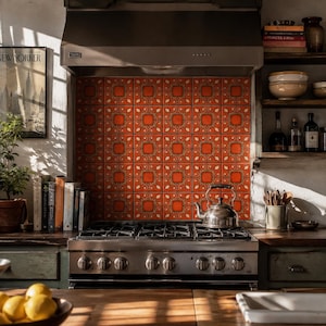 May include: A kitchen backsplash featuring a grid of square tiles with a vibrant orange centre and a decorative cream and brown border pattern. The tiles are installed behind a stainless steel gas range with a matching overhead vent hood.