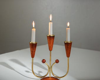 May include: A three-candle candelabra with a mid-century modern design. The candelabra features a wooden base and candle holders, with brass accents and curved arms. Three white candles are lit, casting a warm glow. The overall color scheme is brown, gold, and white.