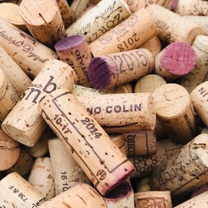 May include: A close-up of a pile of used wine corks. The corks are made of natural cork and have a variety of colors, shapes, and sizes. Some of the corks have writing on them, including dates, names, and logos.